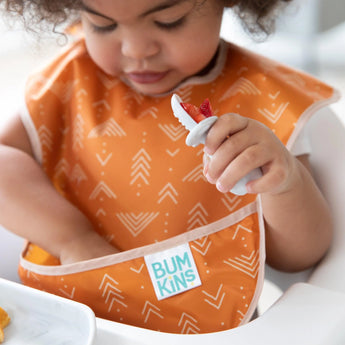 A toddler uses a spoon to eat berries while wearing an orange Bumkins SuperBib® 3 Pack: Desert Boho. Their hand reaches into the pocket.