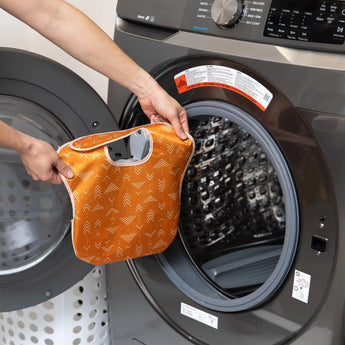 A person holds an orange Bumkins SuperBib® 3 Pack: Desert Boho in front of an open washing machine.