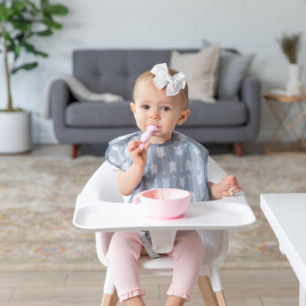 A toddler with a white bow sits in a high chair, wearing a Bumkins SuperBib®: Arrow and eating from a pink bowl with a spoon.