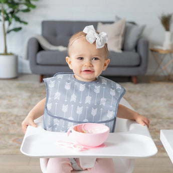 A baby with a bow sits in a high chair in a cozy living room, wearing a Bumkins SuperBib®: Arrow with a pink bowl on the tray.