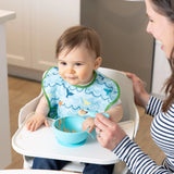A baby in a Bumkins SuperBib® 3 Pack: Rolling With the Waves sits in a high chair beside a smiling woman, with food in a bowl on the tray.