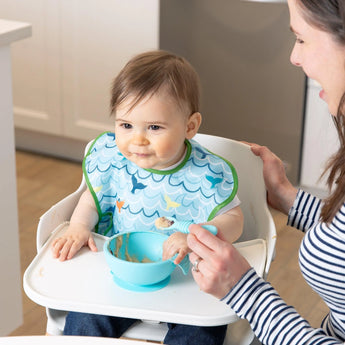 A baby in a Bumkins SuperBib® 3 Pack: Rolling With the Waves sits in a high chair beside a smiling woman, with food in a bowl on the tray.