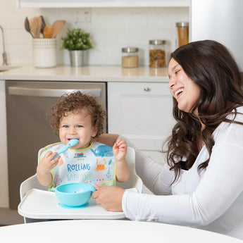 A smiling woman sits by a toddler in a high chair wearing a Bumkins SuperBib® 3 Pack: Rolling With the Waves eating from a blue bowl.