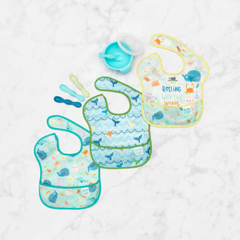 Bumkins SuperBib® 3 Pack: Rolling With the Waves lays flat on a marble surface with a blue bowl & spoon set and 3 toddler spoons nearby.