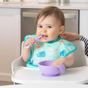 A baby in a high chair wears a Bumkins SuperBib® 3 Pack: Rolling With the Waves, chewing a purple spoon by a purple bowl.