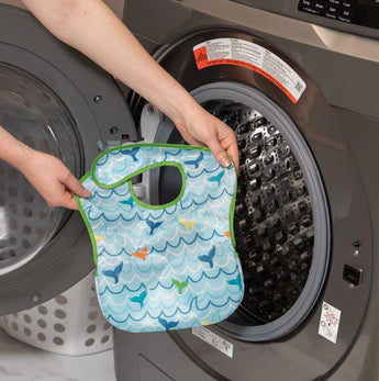 A person holds a Bumkins SuperBib® 3 Pack: Rolling With the Waves in front of an open front-loading washing machine.