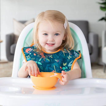 A toddler sits in a high chair, wearing a Bumkins SuperBib® 3 Pack: All Together Now while holding an orange bowl.