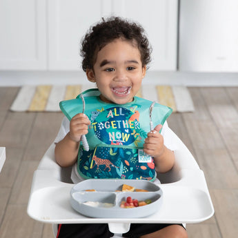 A smiling toddler in a high chair wears a Bumkins SuperBib® 3 Pack: All Together Now, holding utensils near a gray divided plate with food.