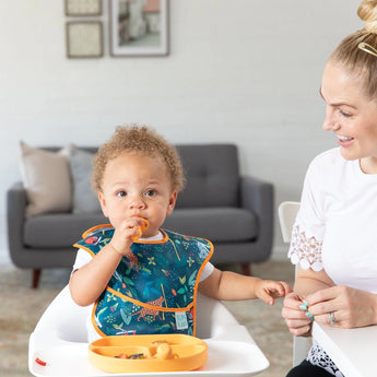 A toddler in a Bumkins SuperBib® 3 Pack: All Together Now eats in a high chair while a smiling woman sits nearby in a cozy living room.