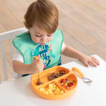 A toddler in a Bumkins SuperBib® 3 Pack: All Together Now enjoys scrambled eggs and fruit with a fork from an orange plate at a table.