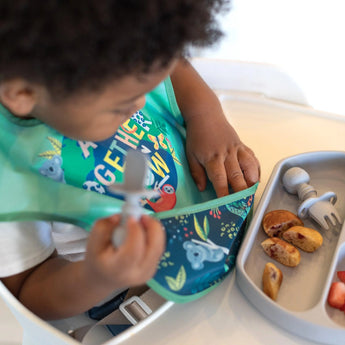 A toddler in a high chair wearing a Bumkins SuperBib® 3 Pack: All Together Now holds a gray spoon while reaching into the bib's pocket.