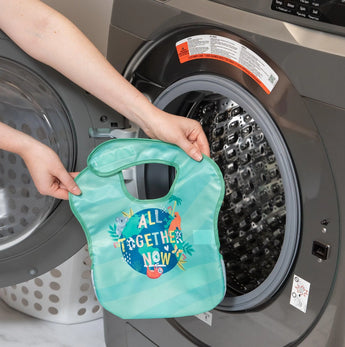 A person holds a Bumkins SuperBib® 3 Pack: All Together Now in front of an open front-loading washing machine.
