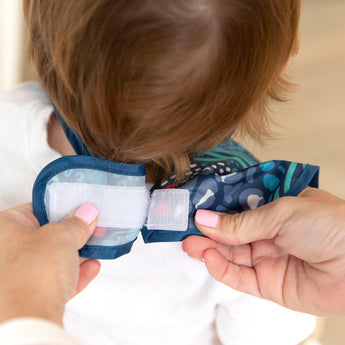 Adult hands fasten the hook-and-loop closure of a Bumkins SuperBib® 3 Pack: All Together Now around a child's neck.