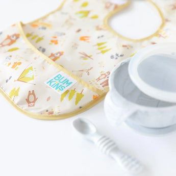 Close-up of a Bumkins SuperBib® 3 Pack: Happy Campers on a white background, with a white marble bowl and spoon in the foreground.