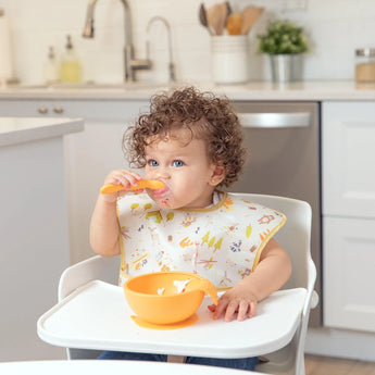 A curly-haired toddler eats with a spoon from an orange bowl, wearing a Bumkins SuperBib® 3 Pack: Happy Campers in a bright kitchen.