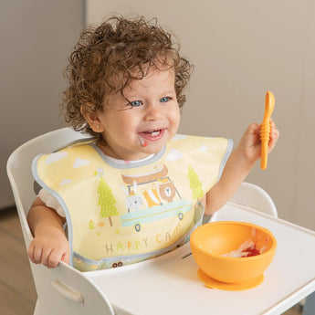 A smiling toddler in a high chair holds an orange spoon by an orange bowl, wearing a yellow Bumkins SuperBib® 3 Pack: Happy Campers.