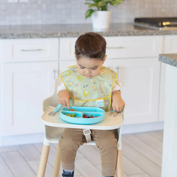 A child in a high chair enjoys food from a blue plate, wearing a green Bumkins SuperBib® 3 Pack: Happy Campers.