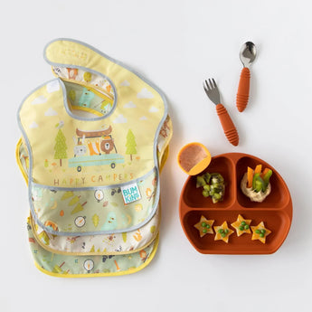 Bumkins SuperBib® 3 Pack: Happy Campers lays on a white background beside a divided plate with food and a set of utensils.