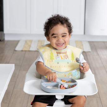 A happy child sits in a high chair, wearing a yellow Bumkins SuperBib® 3 Pack: Happy Campers, holding a fork over a meal in a divided plate.