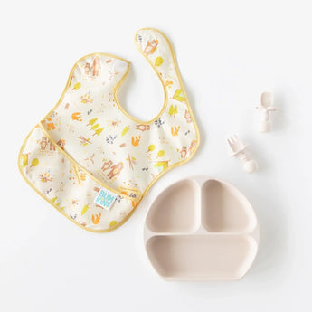 A Bumkins SuperBib® 3 Pack: Happy Campers lays next to a tan divided plate and matching baby utensils on a white background.