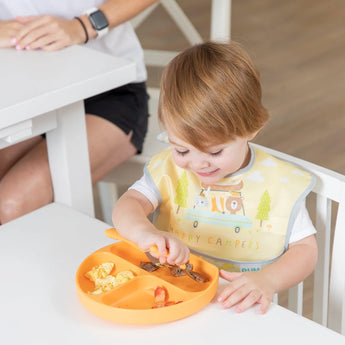 A toddler eats from a orange plate wearing a Bumkins SuperBib® 3 Pack: Happy Campers, sitting at a white table with an adult nearby.