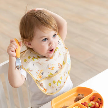 A toddler in a Bumkins SuperBib® 3 Pack: Happy Campers smiles playfully, holding a fork at a table with an orange plate of food.