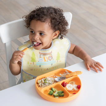 A toddler in a Bumkins SuperBib® 3 Pack: Happy Campers enjoys rice, broccoli, and fruit from an orange sectioned plate at a white table.