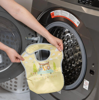 A person holds a yellow Bumkins SuperBib® 3 Pack: Happy Campers in front of an open front-loading washing machine.