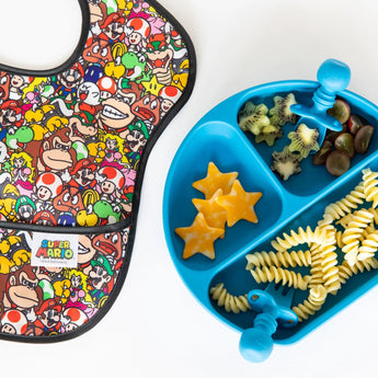 A Bumkins SuperBib® 3 Pack: Super Mario™ Power-Up lays next to a blue plate with cheese, pasta, fruit, and utensils on a white background.