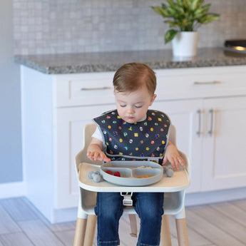 A toddler in a high chair enjoys a meal from a divided plate, wearing a Bumkins SuperBib® 3 Pack: Super Mario™ Power-Up.
