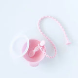A Bumkins Silicone Accessory Tether 2-Pack: Pink is attached to a pink spoon in a pink bowl with a clear lid on a white background.