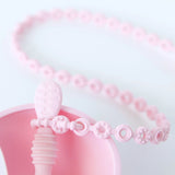 Close-up of a Bumkins Silicone Accessory Tether 2-Pack: Pink attached to a spoon in a bowl on a white background.