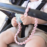 Close-up of a baby in a stroller, holding a pacifier attached to a Bumkins Silicone Accessory Tether 2-Pack: Pink.