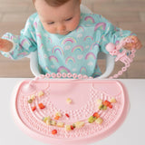 A baby in a rainbow bib eats fruit on a placemat, holding a pink toddler fork attached to a Bumkins Silicone Accessory Tether 2-Pack: Pink.