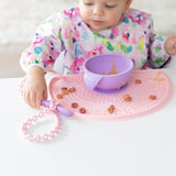 A baby in a bib eats from a purple bowl on a pink mat, holding a purple spoon attached to a Bumkins Silicone Accessory Tether 2-Pack: Pink.