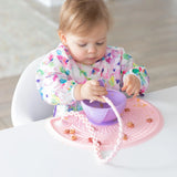 A toddler in a floral bib eats from a bowl on a pink placemat, with a spoon attached to a Bumkins Silicone Accessory Tether 2-Pack: Pink.