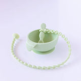 A Bumkins Silicone Accessory Tether 2-Pack: Sage is attached to a green spoon in a green bowl on a white background.