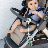 A baby in a stroller joyfully gazes at the camera with a teether on a Bumkins Silicone Accessory Tether 2-Pack: Sage dangling from the side.