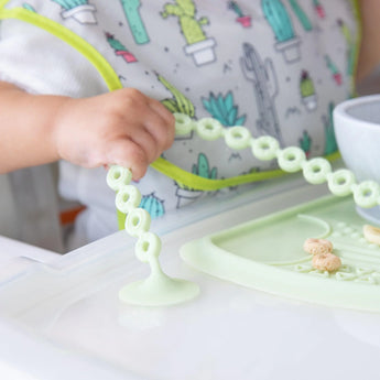 Close-up of a baby wearing a cactus-print bib in a high chair holds a Bumkins Silicone Accessory Tether 2-Pack: Sage attached to the tray.