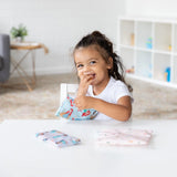 A child beams at a white table with Bumkins Reusable Snack Bag, 3-Pack: Princess Magic, Ariel, and Jasmine in a bright room.