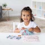A young girl grins at a table, holding Bumkins Reusable Snack Bag, 3-Pack: Princess Magic, Ariel, and Jasmine in a bright room with shelves.