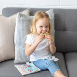 A blonde girl in a white shirt and jeans eats cookies from Bumkins Reusable Snack Bag, 3-Pack: Princess Magic, Ariel, and Jasmine on her lap.