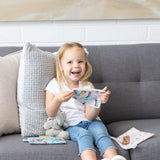 A smiling young girl on a sofa holds Bumkins Reusable Snack Bag, 3-Pack: Princess Magic, Ariel, and Jasmine, a stuffed animal beside her.