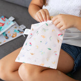 A child on couch opens a Bumkins Reusable Snack Bag, 3-Pack: Princess Magic, Ariel, and Jasmine, with two bags beside her.