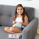 A girl sits on a sofa, holding a Bumkins Reusable Snack Bag, 3-Pack: Princess Magic, Ariel, and Jasmine. Two more bags are beside her.
