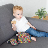 A curly-haired toddler on a gray couch holds a cookie beside Bumkins Reusable Snack Bag, 3-Pack: Super Mario™ Power Up.