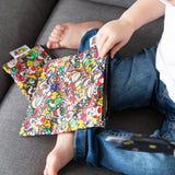 A child in jeans sits on a gray couch, holding a Bumkins Reusable Snack Bag, 3-Pack: Super Mario™ Power Up.