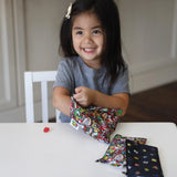 A smiling girl opens a Bumkins Reusable Snack Bag, 3-Pack: Super Mario™ Power Up at a white table.