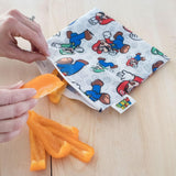 Hands place orange pepper slices into a Bumkins Reusable Snack Bag, Large: Super Mario™ & Luigi on a wooden surface.