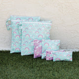 Bumkins Wet Bag: Rainbows sits on grass against a textured beige wall among other matching waterproof bags.